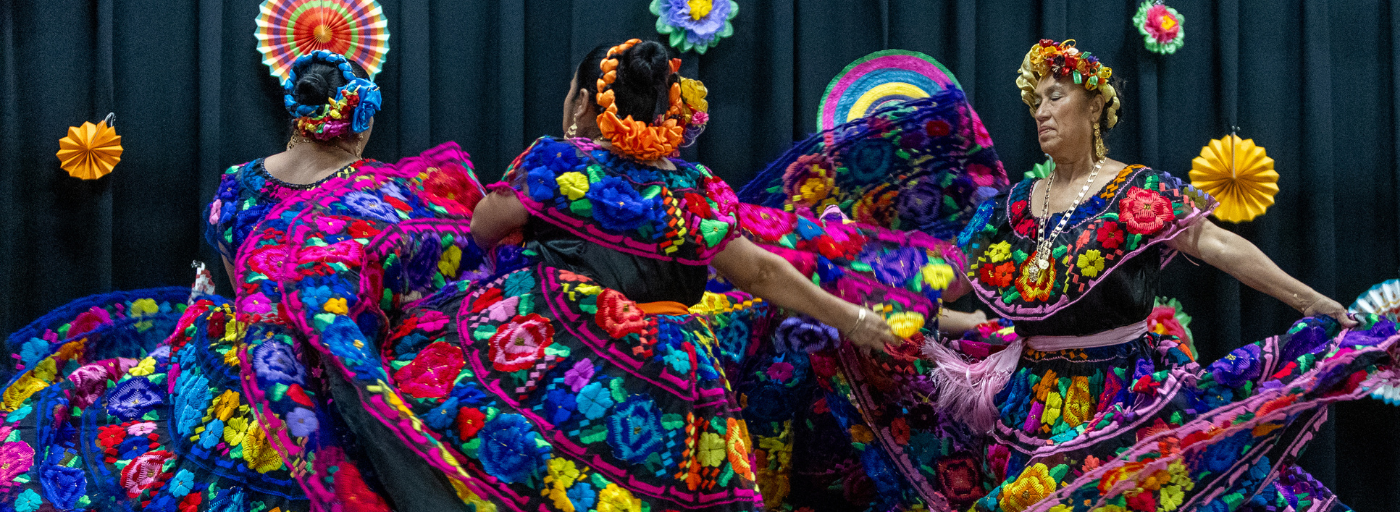 Three adult dancers in vibrant dresses twirl on the stage during the Hispanic Heritage Program.