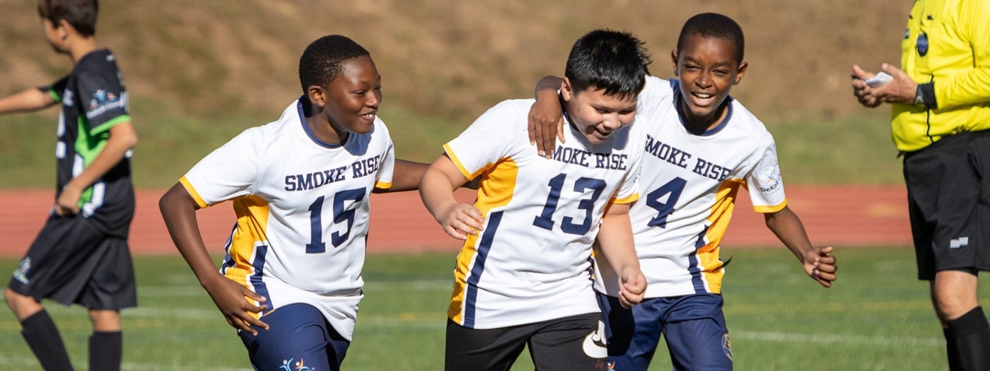 Three students huddled together smiling on the soccer field. 