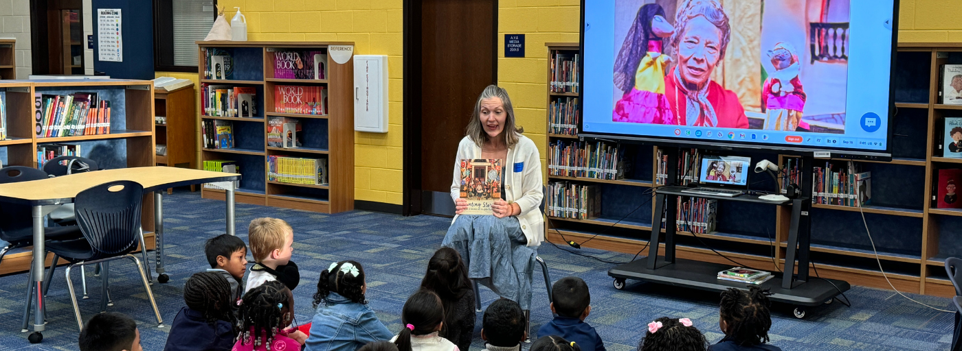 A parent guest reader holding a book with a screen behind her displaying a picture of Pura Belpre. Students are sitting in front listening. 